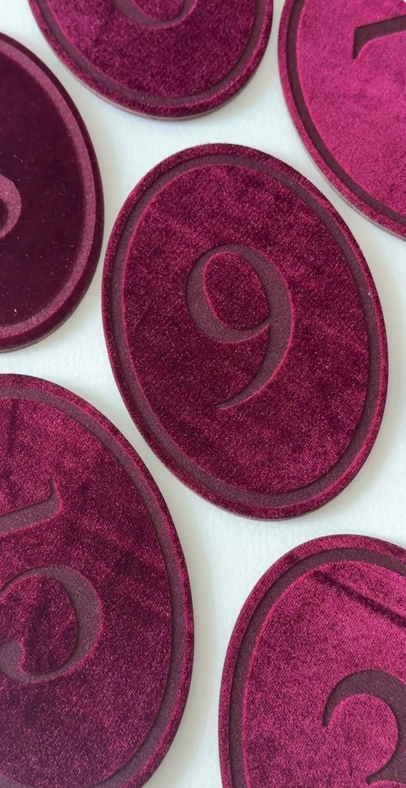 K&ouml;nnte beinhalten: Eine Sammlung ovaler Marker mit einer tiefburgunderroten Samttextur. Jeder Marker zeigt eine einzelne gepr&auml;gte Ziffer in der Mitte, umgeben von einem erhabenen Rand. Die sichtbaren Zahlen umfassen neun, f&uuml;nf, drei und eins.