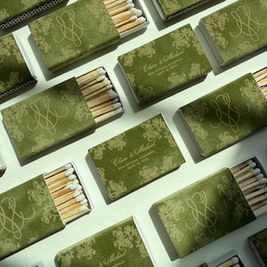 May include: Multiple rectangular matchboxes featuring a green cover with a gold floral pattern and a monogram. Some boxes are open, revealing wooden matches with white tips. Text on the boxes reads: Claire & Michael, New Orleans, Louisiana, April 16, 2026.