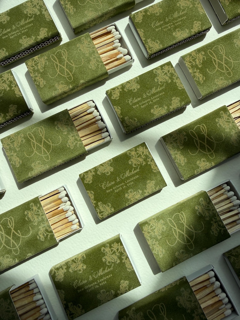 May include: Multiple rectangular matchboxes featuring a green cover with a gold floral pattern and a monogram. Some boxes are open, revealing wooden matches with white tips. Text on the boxes reads: Claire & Michael, New Orleans, Louisiana, April 16, 2026.