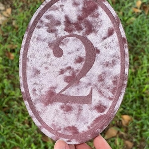 K&ouml;nnte beinhalten: Ein ovaler Aufn&auml;her mit einem Batikmuster in Altrosa und Mauve, in dessen Mitte eine dunkel-mauvefarbene, gestickte Zahl zwei zu sehen ist. Der Aufn&auml;her hat einen gestickten Rand und eine weiche, samtartige Textur auf der gesamten Stoffoberfl&auml;che.