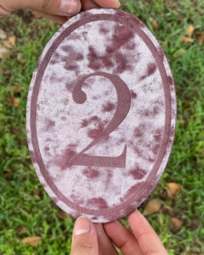K&ouml;nnte beinhalten: Ein ovaler Aufn&auml;her mit einem Batikmuster in Altrosa und Mauve, in dessen Mitte eine dunkel-mauvefarbene, gestickte Zahl zwei zu sehen ist. Der Aufn&auml;her hat einen gestickten Rand und eine weiche, samtartige Textur auf der gesamten Stoffoberfl&auml;che.