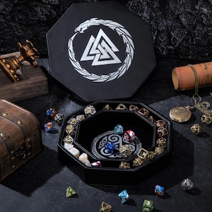 May include: A black octagonal dice box with a white emblem and a matching tray. The tray contains various polyhedral dice in multiple colors, including gold, blue, and green. A treasure chest and other decorative items are also present.