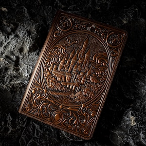 May include: A rectangular notebook with a dark brown, embossed cover featuring a detailed relief of a castle with multiple towers and spires set within a circular frame, surrounded by intricate swirling patterns and a small flying creature.