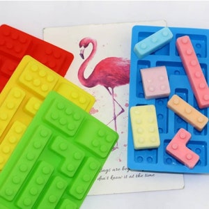 May include: Four silicone moulds for making chocolate or candy in the shape of Lego bricks. The moulds are red, yellow, green, and blue. The blue mould is filled with colourful candy shaped like Lego bricks.