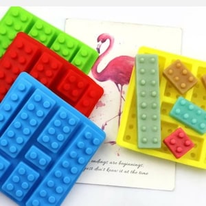 May include: A set of five silicone moulds for making chocolate or candy in the shape of Lego bricks. The moulds are in various colours, including blue, green, red, and yellow. The moulds are on a white surface with a pink flamingo illustration and the text "Endings are beginnings, just don't know it at the time."