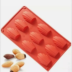 May include: Red silicone mould with nine individual cavities shaped like seashells. The mould is designed for baking madeleine cakes. There are six madeleine cakes on a white surface.