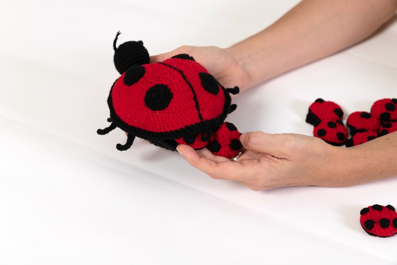 Op de afbeelding: Een gehaakt lieveheersbeestje speelgoed met zwarte poten en antennes, een rood lichaam met zwarte stippen en een kleiner lieveheersbeestje speelgoed bevestigd aan de rug. De lieveheersbeestje speelgoed bevinden zich op een wit oppervlak.