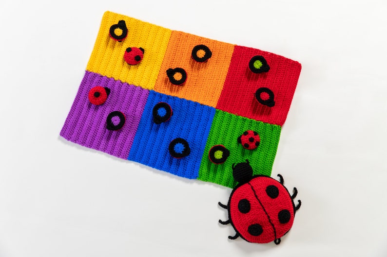 Op de afbeelding: Een kleurrijk gebreid speelkleed met een grote rode lieveheersbeestje in de rechterbenedenhoek. Het kleed is verdeeld in zes vierkanten, elk in een andere kleur: geel, oranje, rood, groen, blauw en paars. Elk vierkant heeft zwarte gebreide cirkels en lieveheersbeestjes.