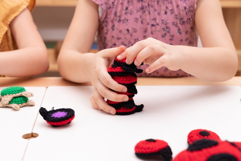 Op de afbeelding: Een kind stapelt gehaakte rode en zwarte lieveheersbeestjes op een witte tafel. De lieveheersbeestjes hebben zwarte stippen en zijn gehaakt met een zwart en rood garen.