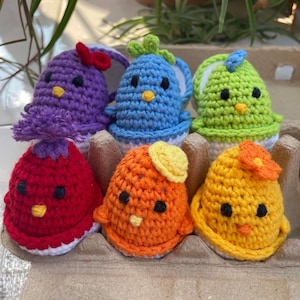 May include: Six crocheted chicks in an egg carton. The chicks are purple, blue, green, red, orange, and yellow. Each chick has a different colored flower or hat.