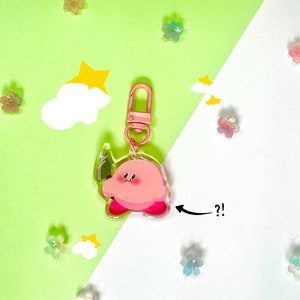 May include: Pink and white cartoon character keychain with a pink and white cloud and a yellow star in the background. The keychain is on a white and green surface with small pastel flowers.