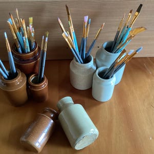 May include: A collection of paintbrushes in various sizes and colours are organised in five small ceramic jars. The jars are a mix of brown and light grey.