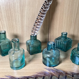 May include: A collection of six small, teal-coloured glass bottles in various shapes, including square and round, displayed on a wooden surface. One bottle holds a decorative feather. The bottles are reminiscent of antique designs.