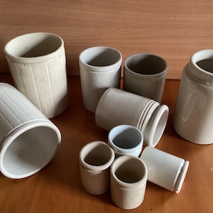 May include: A collection of ten white ceramic jars of varying sizes and shapes. The jars have a smooth, slightly textured surface and are arranged on a wooden surface.