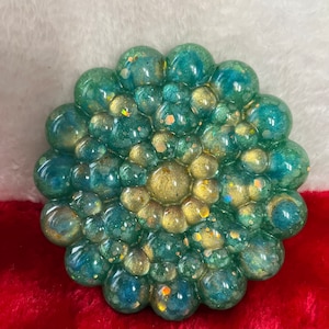 May include: A round, green and gold resin coaster with a raised, circular pattern. The coaster has a shiny, iridescent finish.