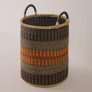 May include: A tall, cylindrical woven basket with two arched handles. The basket features horizontal stripes in black, tan, and orange. The handles are black with tan accents. This decorative basket is suitable for storage or display.