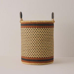 May include: A tall, cylindrical woven basket with two handles. The basket has a repeating pattern of black dots on a tan background. It features bands of black and orange near the top and bottom. The handles are black and tan.