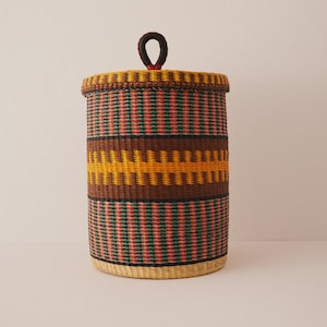 May include: A cylindrical woven basket with a lid and a handle. The basket features horizontal bands of colour, including brown, yellow, green, and pink. The handle is a dark brown loop.