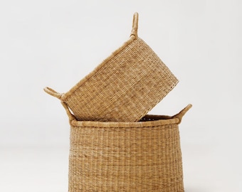 May include: Two woven wicker baskets. One is rectangular with handles, resting atop a larger, round basket with handles. Both are a natural tan color, with a textured weave pattern. The baskets are empty and set against a white background.