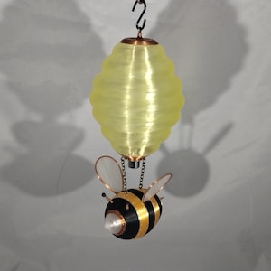 May include: A yellow, honeycomb-shaped paper lantern hangs from a chain, suspended above a black and yellow bumblebee figurine. The bumblebee has clear wings and a copper-colored body.