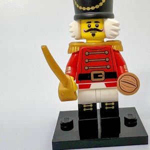 Peut inclure: Une figurine Lego de casse-noisette soldat. Il porte un uniforme rouge et or avec un chapeau noir et une bordure en fourrure blanche. Il tient une épée dorée et un casse-noisette brun.