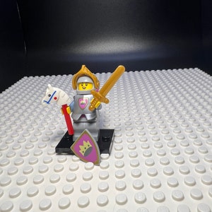 Peut inclure: Une figurine Lego de chevalier féminin portant une armure argentée et rose, une couronne dorée et tenant une épée et un cheval en jouet. La figurine est debout sur une base noire avec un bouclier violet orné d'une couronne dorée.
