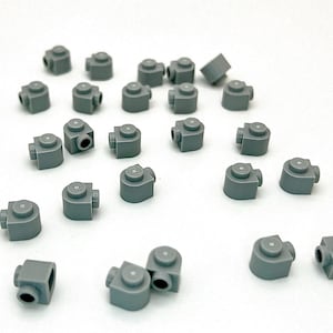 May include: A collection of small, gray plastic toy heads scattered on a white surface. Each head has a cylindrical neck and a circular top. The heads are designed for building block figures.