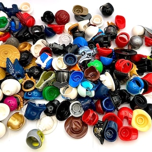 May include: A collection of colorful plastic toy helmets and headgear in various shapes and sizes. The assortment includes hats, helmets, and head coverings in red, black, blue, yellow, and white. Some pieces have unique designs, such as a cowboy hat and a Viking helmet.