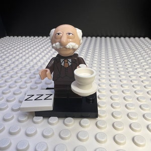 May include: A brown and white Lego minifigure of a man with a mustache and a cup in his hand. The minifigure is standing on a black base with a white plate that says "ZZZ" on it.