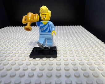 May include: A Lego figure of a figure skater holding a gold trophy. The figure is wearing a blue and white skating outfit and has blonde hair. The figure is standing on a black base.