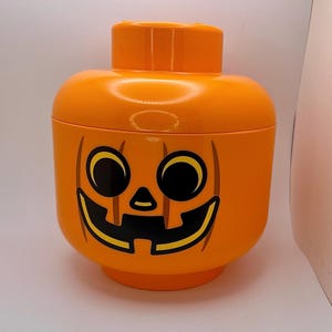 May include: An orange plastic container shaped like a large head, with a jack-o'-lantern face. The container has a lid and a round base. The jack-o'-lantern face is black and yellow, with large eyes and a smiling mouth. A fun Halloween decoration.
