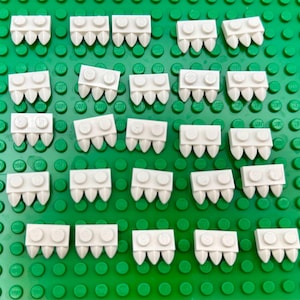 May include: White Lego bricks with a tooth-like design are arranged on a green Lego baseplate. The bricks have two circular studs on top and three pointed teeth on the bottom. The image is a top-down view.
