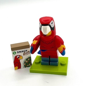 May include: A red, yellow, and blue parrot figure with a white beak stands on a green base. Beside it is a small snack box with a parrot illustration and the words "SNACK BOX". The figure is a toy.