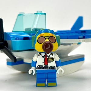 May include: A Lego seaplane with a blue and white design, featuring a black propeller and a pilot minifigure. The pilot wears a yellow head with goggles and a brown nose, a white shirt, blue pants, and the letters "DAL" on the shirt.