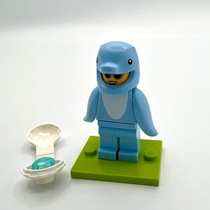 May include: A light blue dolphin costume minifigure with a smiling face, standing on a green base. A white clam shell with a blue gem is next to the figure. The figure has a light blue dolphin head.