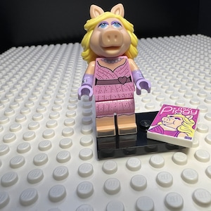 May include: A pink and purple Miss Piggy Lego minifigure wearing a pink dress and purple gloves. The minifigure is standing on a black base with a small magazine featuring Miss Piggy on the cover. The magazine is titled "Miss Piggy".
