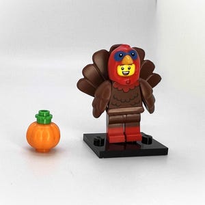 Peut inclure: Une figurine Lego habillée en dinde avec un costume brun et rouge et une citrouille. La figurine est debout sur une base noire.