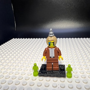 May include: A brown Lego minifigure with a yellow head and a silver antenna on top. The minifigure has a blue and black screen for a face and is standing on a black base with silver stars. Two green Lego minifigures are on either side of the base.