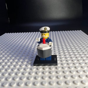 Peut inclure: Une figurine Lego de capitaine portant un uniforme bleu, un chapeau blanc avec une ancre et une écharpe rouge. Le capitaine est debout sur une base noire avec un volant de navire blanc.