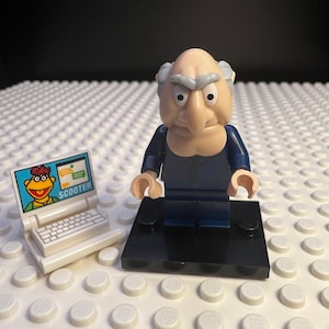 May include: A Lego figure of a stern-faced man in a dark blue suit stands on a black base. Beside him is a small white laptop with a screen featuring a cartoon character and the word "SCOOTER". The background is a white Lego baseplate.