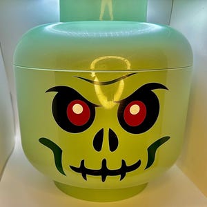 May include: A light green, large plastic container shaped like a Lego head with a zombie face design. The zombie face has black eyebrows, eye sockets, and mouth, with red eyes. The container has a lid and is likely for storage.