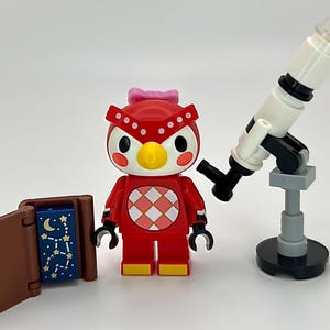 May include: A red owl figurine with a pink bow, a white and black telescope, and a brown book with a blue starry page. The owl has a yellow beak and a diamond pattern on its chest. The telescope is approximately 15 cm tall.