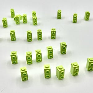 May include: A collection of small, lime green building blocks scattered on a white surface. Each block has a rectangular shape with two circular studs on top and two on the side, typical of construction toys.