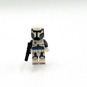 May include: A white and gray plastic toy soldier minifigure featuring a detailed helmet with a black visor, a torso with blue and black accents, and white legs with gray detailing. The figure holds a small black plastic blaster accessory.