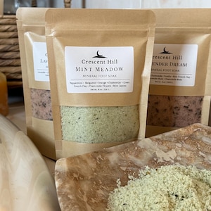 Puede incluir: Se muestran tres bolsas de papel marrón de baño de pies mineral. La bolsa central está etiquetada como "Crescent Hill Mint Meadow" y enumera los ingredientes. Las bolsas contienen sales minerales verdes y rosas. Un cuenco de mármol contiene un montón de baño de pies verde.
