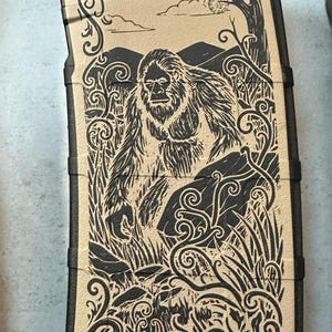 May include: Tan and black magazine with a detailed illustration of a Bigfoot in a forest scene. The design includes mountains, trees, and decorative scrollwork. The text "5.56 223" is visible.