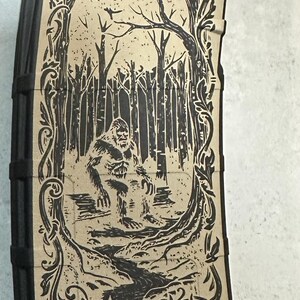 May include: Black magazine with a tan graphic of a Bigfoot in a forest scene. The design is framed by ornate scrollwork. The Bigfoot is depicted standing near a stream in a forest.