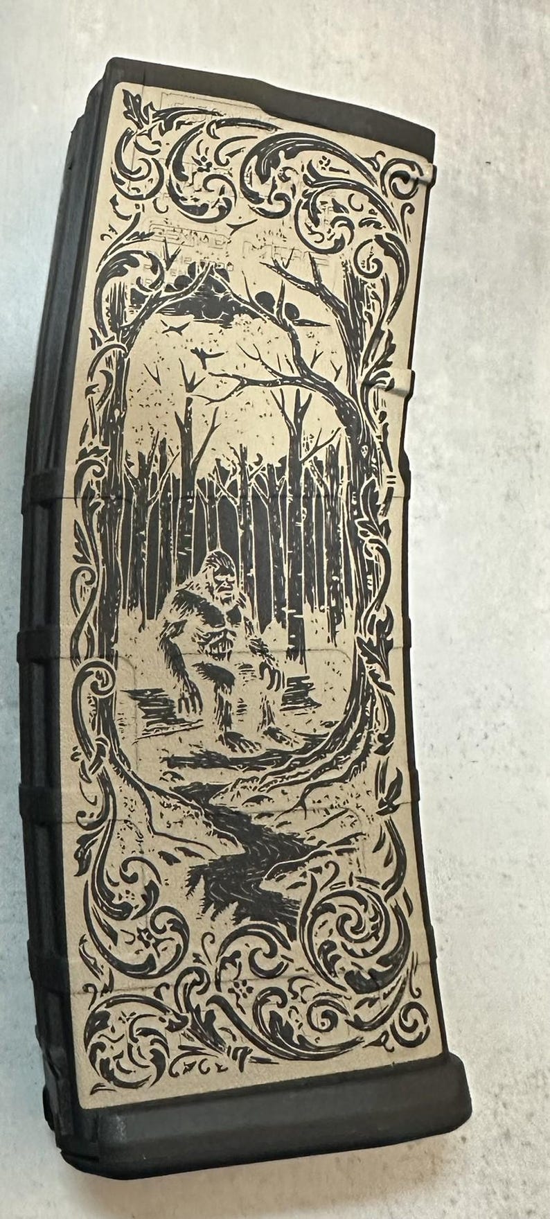 May include: Black magazine with a tan graphic of a Bigfoot in a forest scene. The design is framed by ornate scrollwork. The Bigfoot is depicted standing near a stream in a forest.