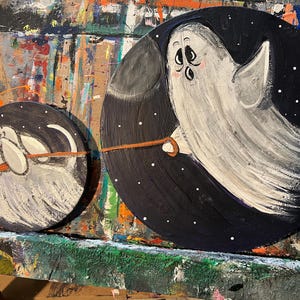 Könnte beinhalten: Zwei runde, bemalte Halloween-Dekorationen. Eine zeigt einen weißen Geist mit einer Leine, die andere ein weißes, hundeähnliches Wesen. Beide sind vor einem dunkelblauen Hintergrund mit weißen Sternen. Der Geist hält die Leine.