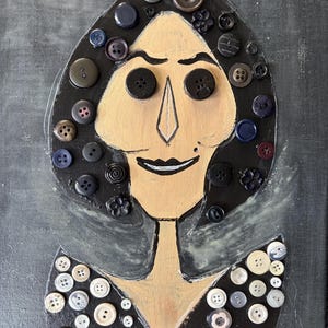 Könnte beinhalten: Ein Mixed-Media-Kunstwerk auf Leinwand, das ein stilisiertes Porträt einer Figur mit bemaltem beigen Gesicht und schwarzem Haar zeigt. Das Haar und die Kleidung sind mit zahlreichen schwarzen, weißen und grauen Knöpfen verschiedener Größen verziert.
